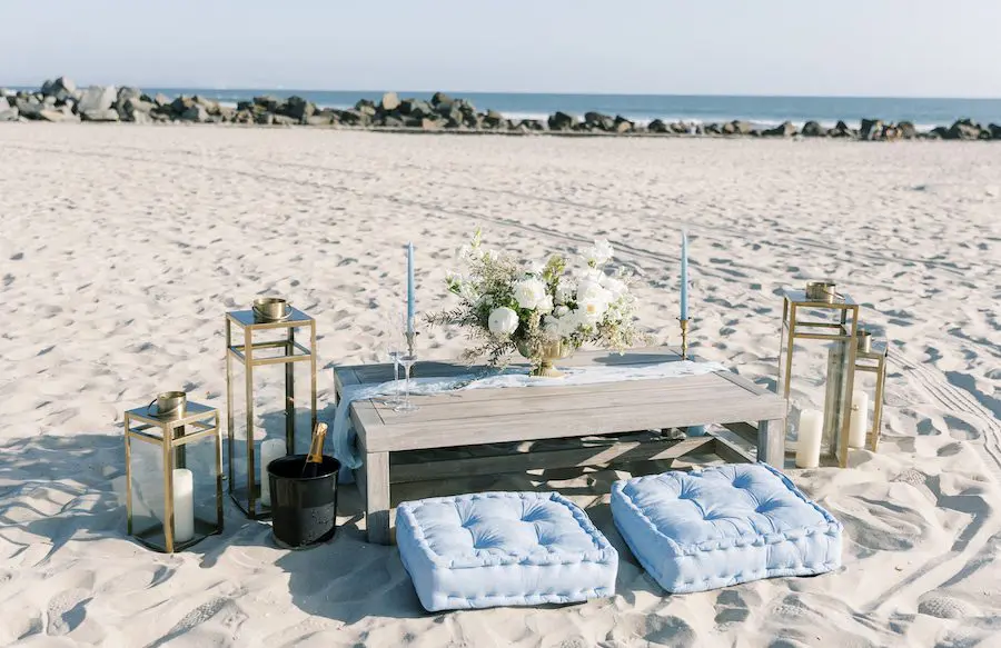 wedding proposal setup on the beach with wooden table, champagne, and seating cushions