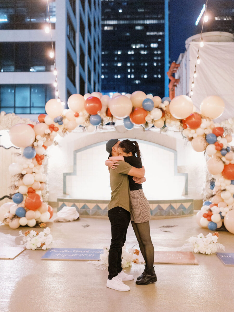 couple hugging blue orange proposal blue orange balloons balloon arch proposal floral balloon arch game night proposal game board proposal rooftop proposal los angeles proposal los angeles rooftop proposal california proposal