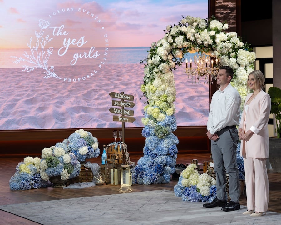 Riley and Joe Canty Present their business to the Sharks on Shark Tank for the opportunity to invest in their proposal planning business. A blue floral arch stands on set with a hanging chandelier, with ocean inspired decor to make this proposal set up truly one of a kind!