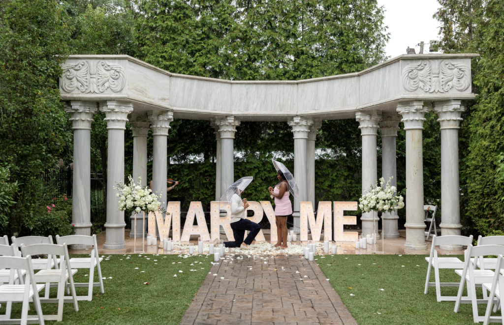 Romantic wedding scene of Sam and Lola under a classical pavilion with large "MARRY ME" letters and.