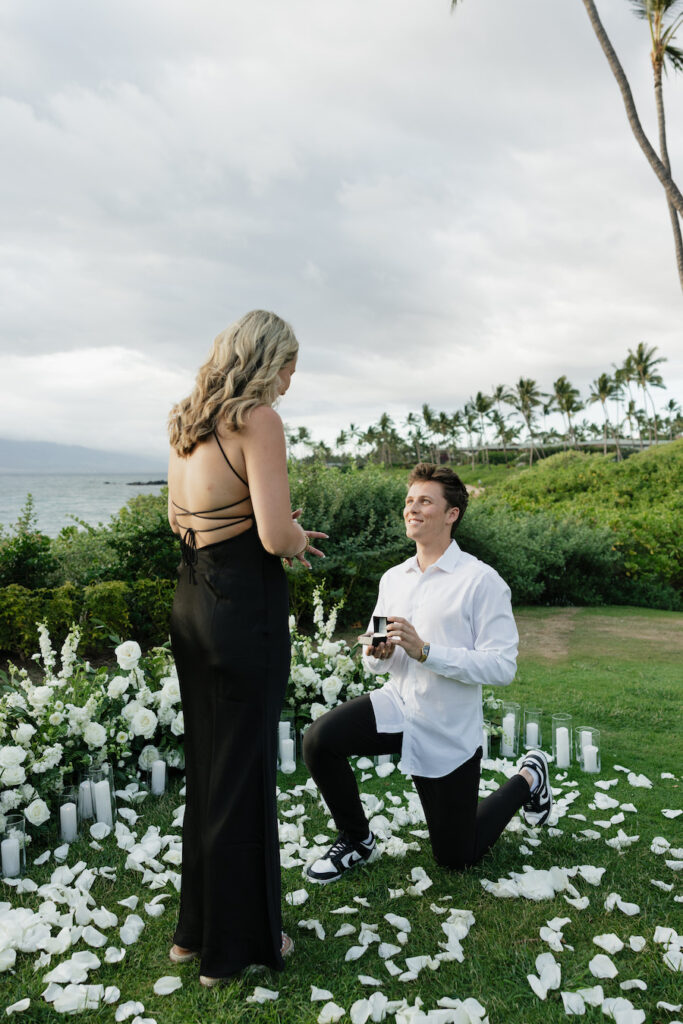 Man proposes to woman on beach with ocean view and floral decor.