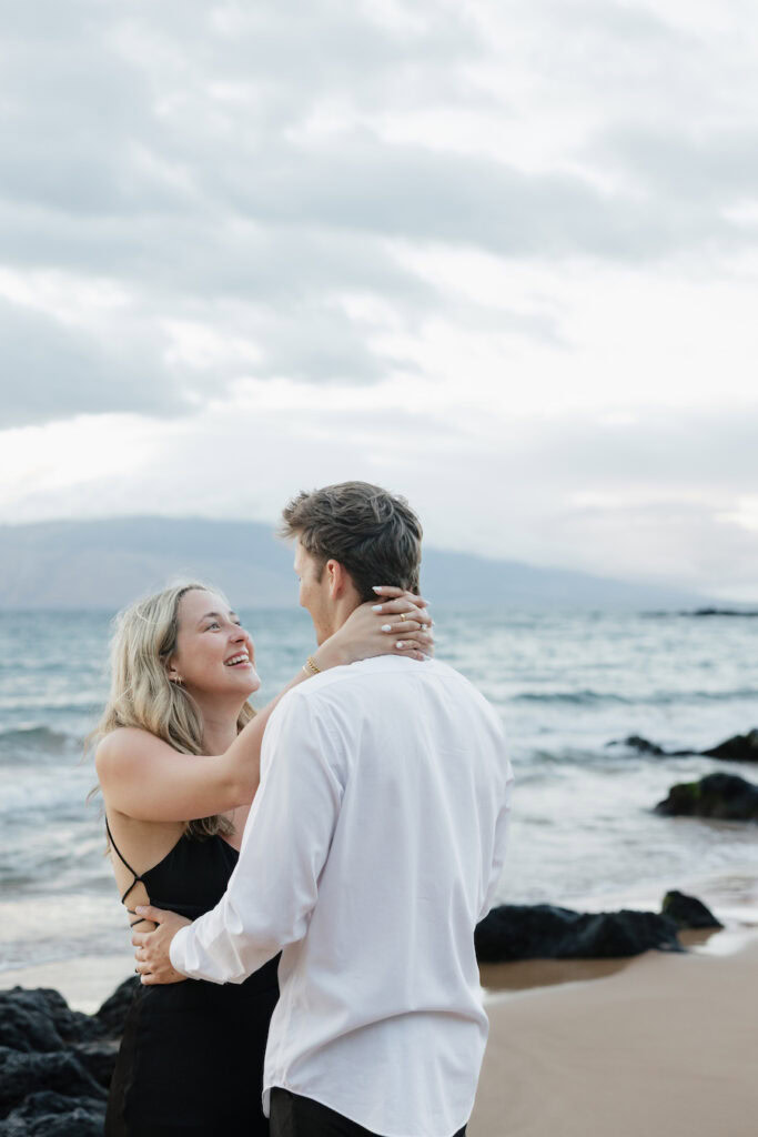 Couple embracing on the beach with ocean and cloudy sky in the background.