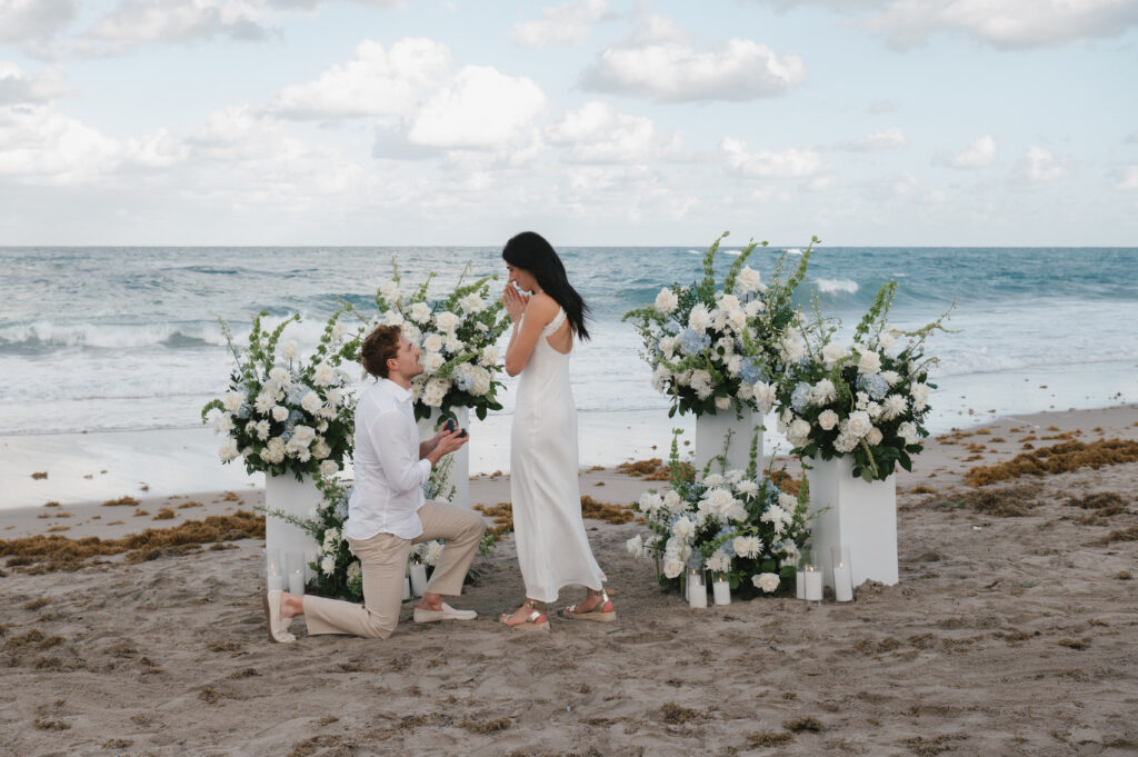 Proposal on the beach with a woman and man surrounded by white flowers and ocean backdrop.