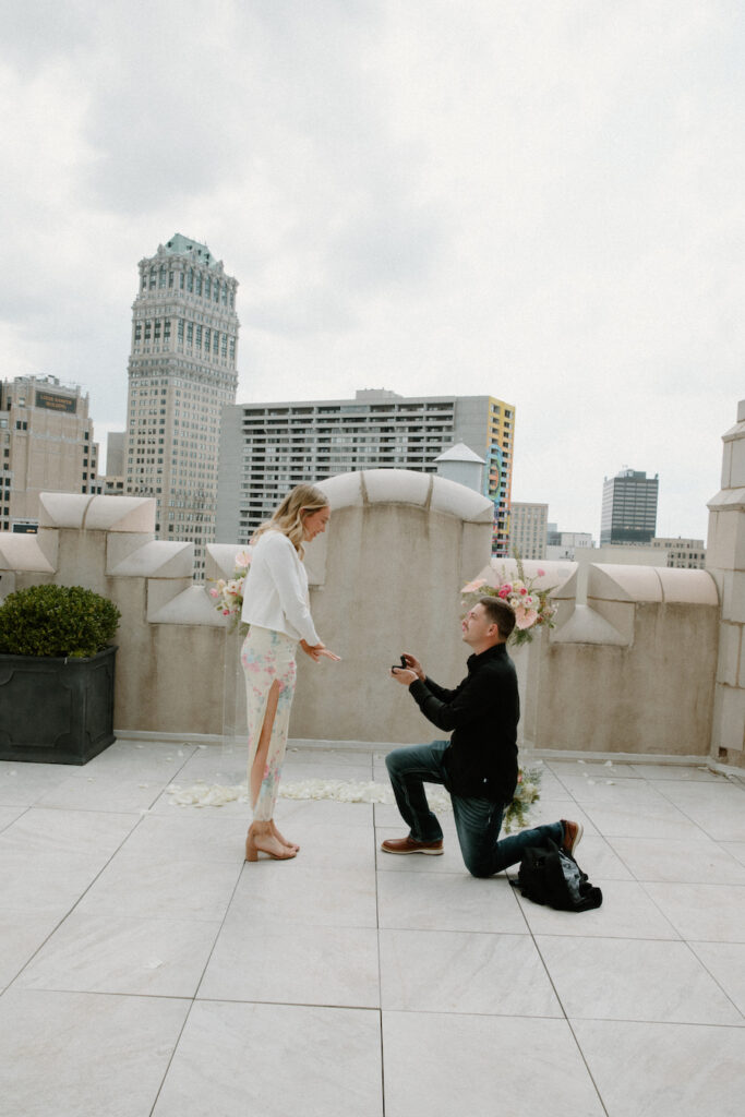 Proposal on rooftop with woman in white dress and man kneeling with ring.