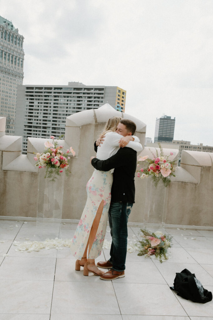 Bride and groom sharing a heartfelt hug on rooftop with city skyline backdrop moments after their proposal.