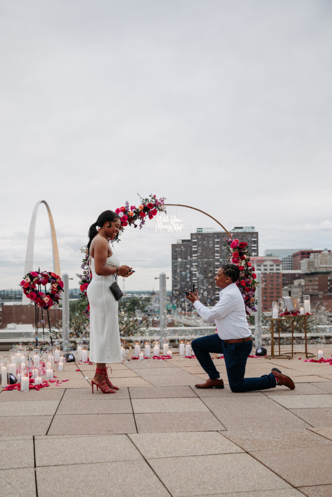 A woman in white dress and red heels is proposed to by a man kneeling with a ring.