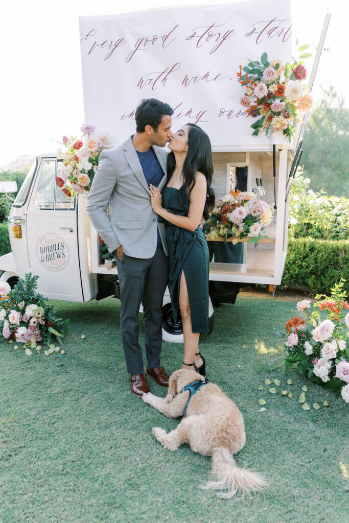 Newly engaged couple kissing with their golden retriever beside a custom calligraphy sign