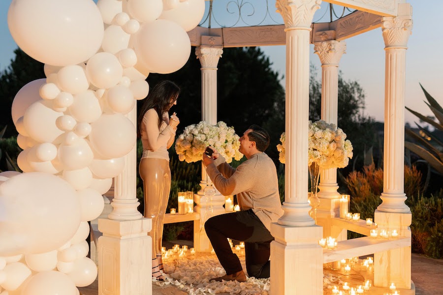 Man on one knee proposing inside a white gazebo with balloons and candles