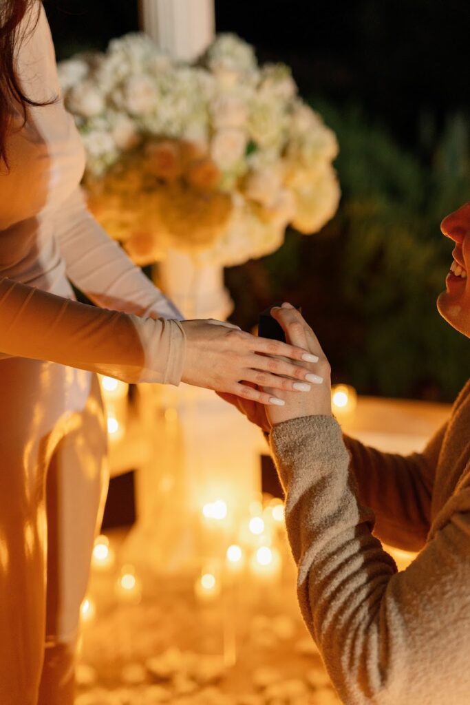 Man on one knee holding her hand surrounded by candlelight and white florals