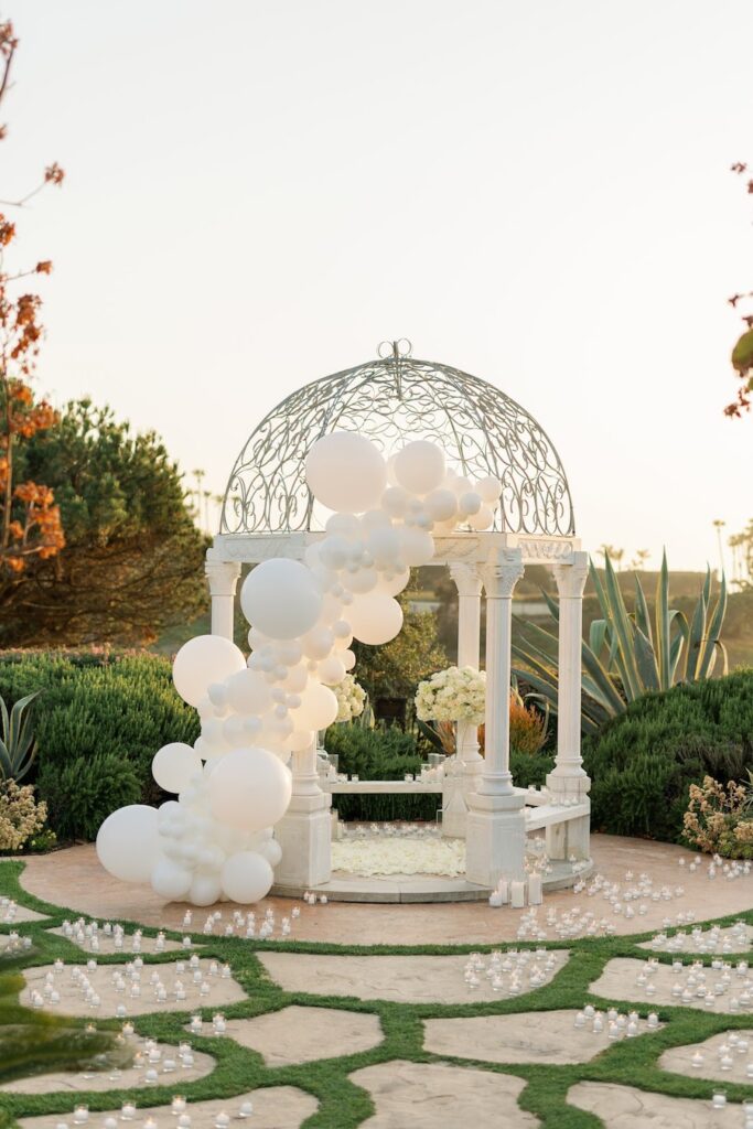 White gazebo proposal setup in Orange County with an ivory balloon garland and candles on the lawn