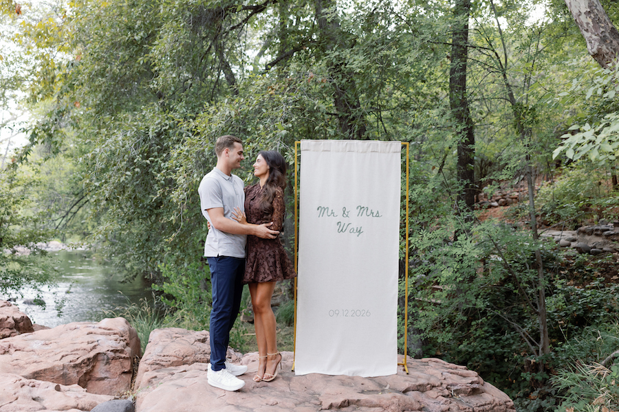 Couple embracing beside a custom welcome banner by the creek