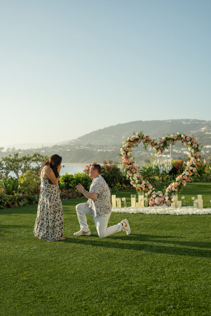 He drops to one knee in front of the heart-shaped floral arch on the oceanfront lawn